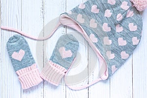 Children`s winter accessories, warm knitted mittens and hat - gray with pink hearts on white wooden background.