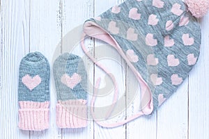 Children`s winter accessories, warm knitted mittens and hat - gray with pink hearts on white wooden background.