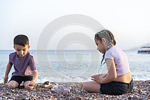 Children show each other sea pebbles. Two kids sitting on sea beach in summer day.