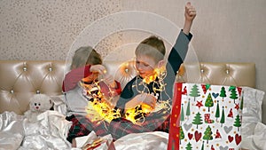 Children sit on the bed, took out a Christmas garland from a beautiful box and play with it. They look at the lights