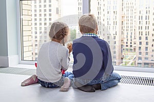 children sit on floor by window and admire view during quarantine. little blond boy and girl with pigtails looking at something