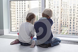 children sit on floor by window and admire view during quarantine. little blond boy and girl with pigtails looking at something