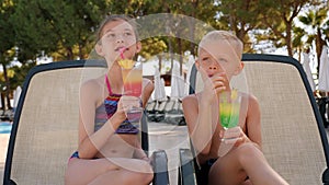Children are sitting on a chaise longue by the pool with bright cocktails.
