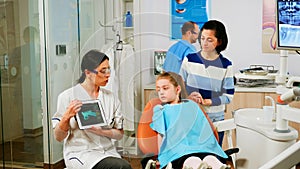 Children sitting on stomatological chair listening pediatric doctor looking on tablet