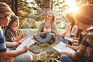 Children and smiling female teacher sitting in circle on grass in forest and talking about nature