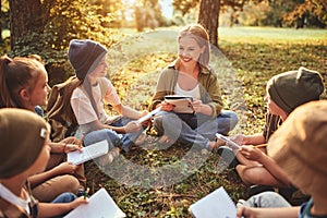 Children and smiling female teacher sitting in circle on grass in forest and talking about nature