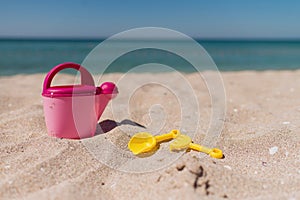 Children toys on a sand beach with blue background. A pink toy watering can with yellow shovel and rake.