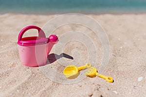 Children toys on a sand beach with blue ocean background. A pink toy watering can with yellow shovel and rake. Vacation with child