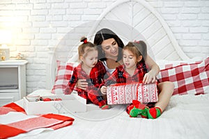 Children unwrap Christmas gifts with their mother sitting in bed in the bedroom