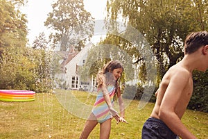 Children Wearing Swimming Costumes Having Fun In Garden Playing In Water From Garden Sprinkler