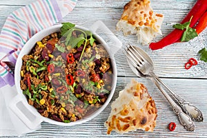 Chili con carne in a white casserole on wooden table with fresh vegetables and bread.