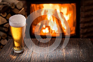 Chilled glass of light beer on the wooden table. Fireplace at the background