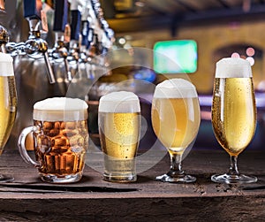 Chilled mugs and glasses of beer on the old wooden table. Pub interior and bar counter with beer taps at the background.