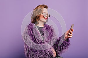 Chilling girl with dark-brown hair wears soft brigth fur coat holding smartphone. Elegant young woman in sunglasses and