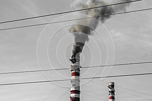 The chimney of a thermal power plant that generates dark smoke, against a dark smoky sky, with high-voltage lines around it