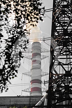 The chimney of a thermal power plant that generates white smoke, against a dark smoky sky, with high-voltage lines around it