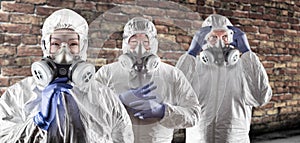 Chinese and Caucasian Woman and Men In Gas Masks, Goggles and Hazmat Suites Against Brick Background photo