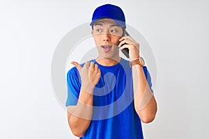 Chinese deliveryman wearing cap talking on the smartphone over isolated white background pointing and showing with thumb up to the