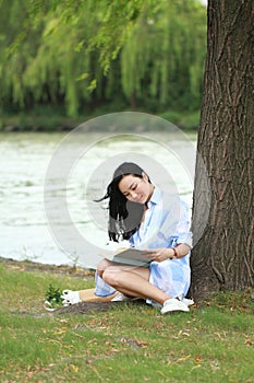 Chinese Girl reading a book under tree. Blonde beautiful young woman with book sit on the grass.