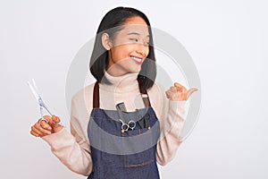 Chinese hairdresser woman wearing apron holding scissors over isolated white background pointing and showing with thumb up to the