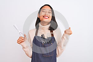 Chinese hairdresser woman wearing apron holding scissors over isolated white background screaming proud and celebrating victory