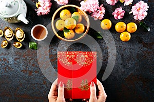 Chinese new year festival decorations. Woman hand holding pow or red packet, orange and gold ingots on a black stone background.