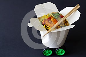 Chinese noodles with shrimps and vegetables in take-out box with car wheels. Creative chinese food delivery concept
