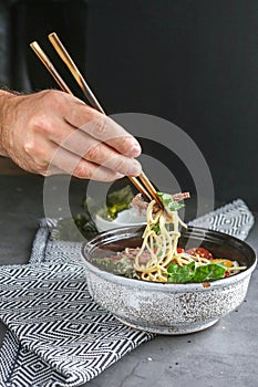 Chinese soup with tomatoes, beef, noodles and herbs on a gray table. the guy picks up macaroni and meat from the plate with wooden