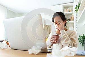 Chinese woman feel sick working from home office. Asian female blowing nose with tissue holding cup of herbal tea.