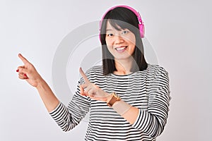 Chinese woman listening to music using pink headphones over isolated white background smiling and looking at the camera pointing