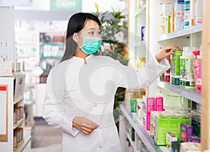 Chinese woman pharmacist in protective facial mask keeps track of drugs in interior of pharmacy
