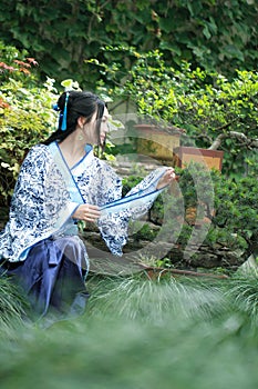 Chinese woman in traditional Blue and white Hanfu dress sit next to bonsai