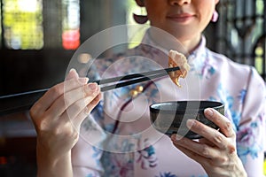 Chinese woman in traditional cheongsam or qipao dress using chopsticks in Chinese restaurant. Fashion woman eating a