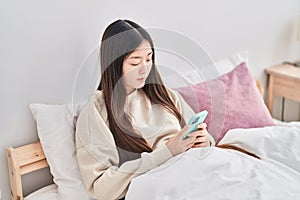 Chinese woman using smartphone sitting on bed at bedroom