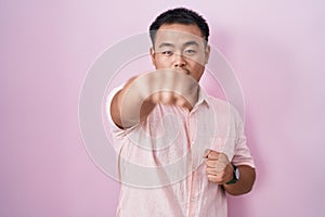 Chinese young man standing over pink background punching fist to fight, aggressive and angry attack, threat and violence