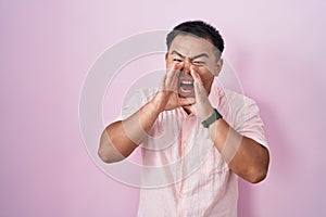 Chinese young man standing over pink background shouting angry out loud with hands over mouth