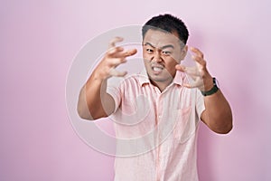 Chinese young man standing over pink background shouting frustrated with rage, hands trying to strangle, yelling mad