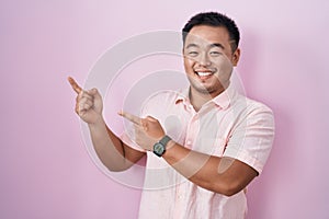 Chinese young man standing over pink background smiling and looking at the camera pointing with two hands and fingers to the side