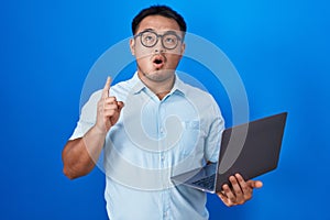 Chinese young man using computer laptop amazed and surprised looking up and pointing with fingers and raised arms