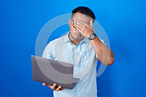 Chinese young man using computer laptop covering eyes with hand, looking serious and sad