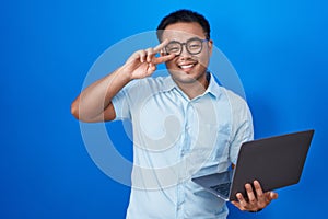 Chinese young man using computer laptop doing peace symbol with fingers over face, smiling cheerful showing victory