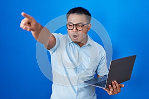 Chinese young man using computer laptop pointing with finger surprised ahead, open mouth amazed expression, something on the front