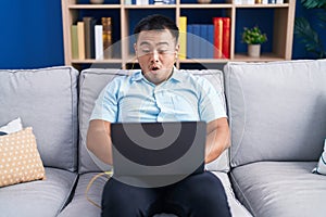 Chinese young man using computer laptop sitting on the sofa scared and amazed with open mouth for surprise, disbelief face