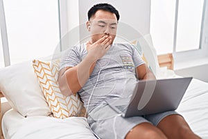 Chinese young man using laptop sitting on the bed covering mouth with hand, shocked and afraid for mistake