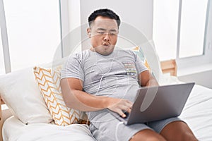 Chinese young man using laptop sitting on the bed scared and amazed with open mouth for surprise, disbelief face