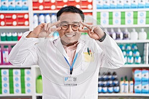 Chinese young man working at pharmacy drugstore doing peace symbol with fingers over face, smiling cheerful showing victory