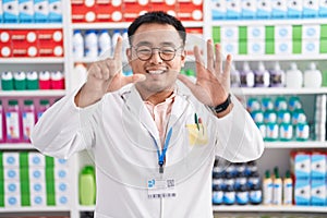 Chinese young man working at pharmacy drugstore showing and pointing up with fingers number seven while smiling confident and
