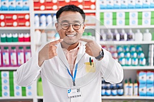 Chinese young man working at pharmacy drugstore smiling cheerful showing and pointing with fingers teeth and mouth
