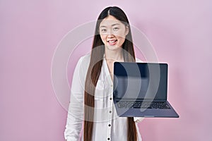 Chinese young woman holding laptop showing screen looking positive and happy standing and smiling with a confident smile showing