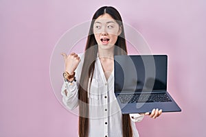 Chinese young woman holding laptop showing screen pointing thumb up to the side smiling happy with open mouth